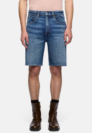 Mężczyzna w niebieskich dżinsowych szortach, czarnych skarpetkach i błyszczących brązowych botkach, stojący na jednolitym białym tle.