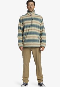 Pull en polaire beige avec des rayures vertes et bleues, doté d'un col montant, d'une fermeture éclair mi-zip et de poches latérales ; associé à un pantalon beige.