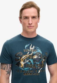 Camiseta de manga corta en color azul verdoso, confeccionada en algodón, con un gráfico de peces koi y texto en dorado y azul, cuello redondo y corte estándar.