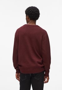 Maglione in maglia borgogna con scollatura rotonda e polsini a coste. Vestibilità ampia con una texture liscia. Pantaloni neri visibili nella parte inferiore.