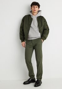 Νεαρός άνδρας με μπουφάν bomber σε ελιά πράσινο χρώμα, παντελόνια στο ίδιο χρώμα, γκρι φούτερ με κουκούλα και μαύρα μοκασίνια, στέκεται δίπλα σε άσπρο τοίχο με τα χέρια στις τσέπες.