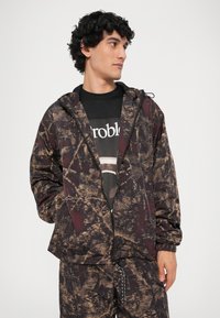 Giacca leggera con cappuccio in un motivo astratto marrone scuro e nero, con chiusura a zip e polsini elasticizzati. Abbinata a una t-shirt nera con grafica.