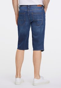 Pantalones cortos de mezclilla en un lavado azul oscuro con un corte recto, dos bolsillos traseros y un ligero deshilachado en el dobladillo, combinados con zapatillas blancas.