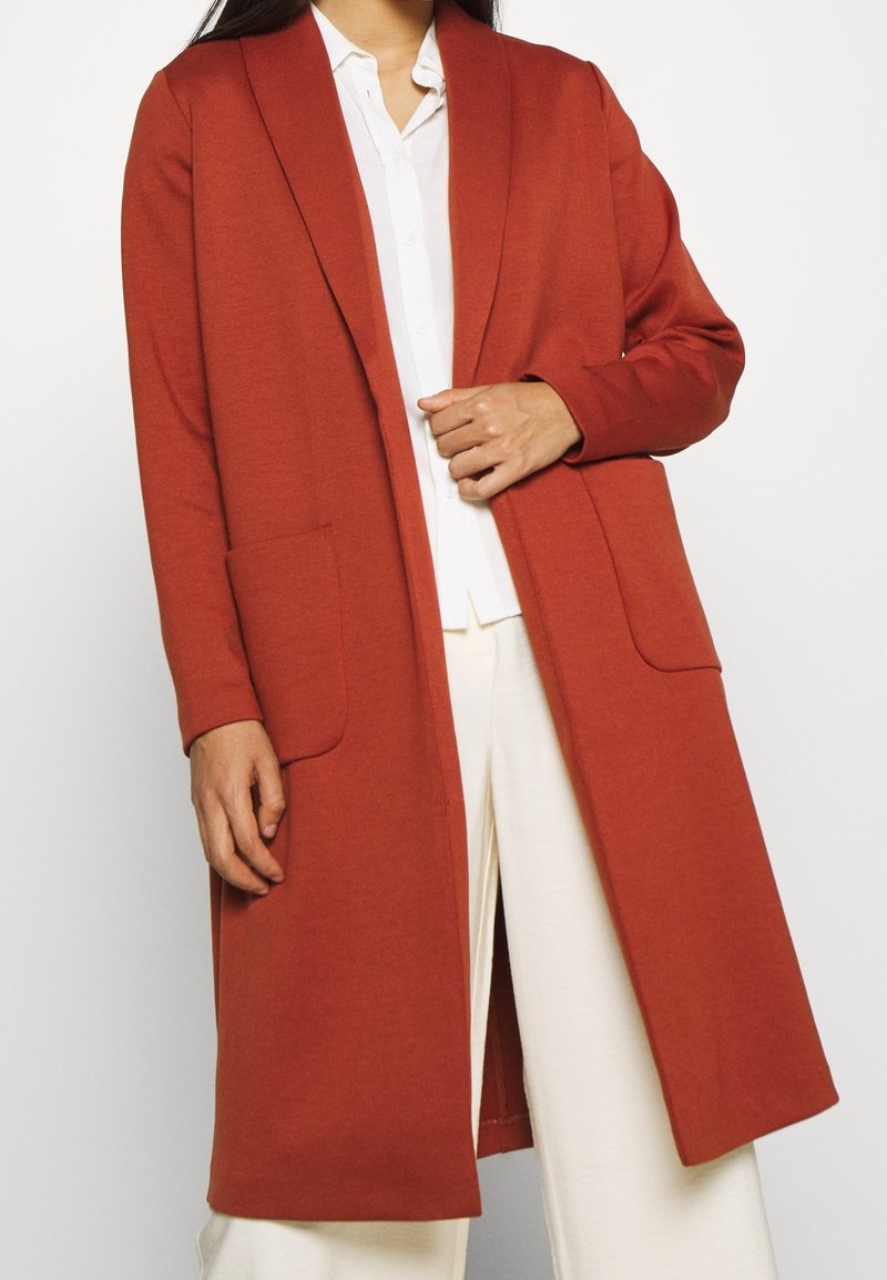 Langer, offener Cardigan in Terrakotta, aus weichem Material. Verfügt über große Fronttaschen und eine lässige Passform. Kombiniert mit einem weißen Hemd.