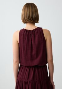 Top senza maniche color bordeaux con scollo arricciato, tessuto testurizzato e chiusura con bottone sul retro. Silhouette fluida con vestibilità morbida.