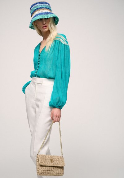 Bluză turquoise cu mânecă lungă și detaliu cu nasturi, asortată cu pantaloni albi, cu talie înaltă, și o poșetă țesută bej cu lanț auriu. pălărie tip bucket cu dungi albastre.