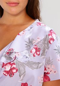 Vaaleanlaventelin värinen paita, jossa on pinkkejä ja harmaita kukkakuoseja. Varustettu röyhelökauluksella ja pehmeällä, sileällä kankaan tekstuurilla.