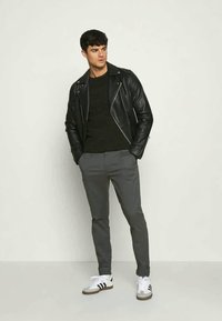 Giacca da biker in pelle nera, maglione a maglia scura, pantaloni grigi slim-fit e sneakers bianche con strisce nere. Texture lisce e design aderente.