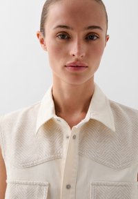 Kermanen hihaton paita, jossa on herringbone-kudos yläosassa, suuret rintataskut ja viistot kaulukset; päällimmäiset napit näkyvillä.