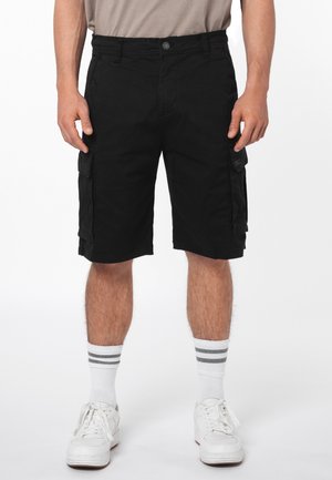 Mann trägt schwarze Cargoshorts, weiße Socken mit grauen Streifen und weiße Sneaker, steht vor einem weißen Hintergrund.
