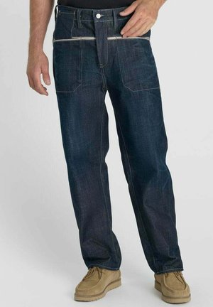 Man draagt donkerblauwe denim jeans met witte ritszakken aan de voorkant en beige suède schoenen tegen een effen achtergrond.