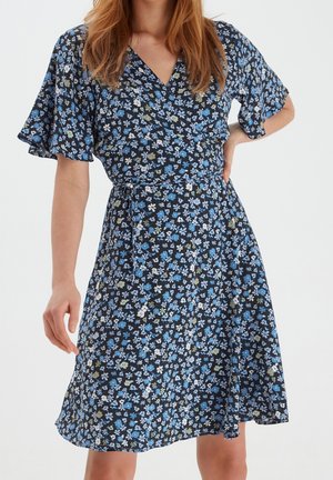 Frau trägt ein knielanges marineblaues Wickelkleid mit blauen, weißen und grünen Blumenmustern und Schmetterlingsärmeln, posiert mit der Hand in der Hüfte.