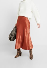 Jupe midi en satin orange rouille, pull en maille blanc surdimensionné, sac à main en imprimé serpent, et bottines à talon bloc.
