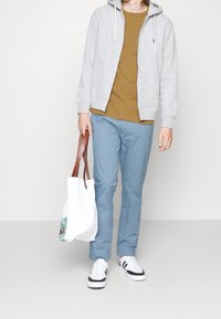 Šedá mikina na zip přes hnědé tričko, modré kalhoty a bílé tenisky s černými akcenty. Béžová a hnědá taška přes rameno s květinovým vzorem.