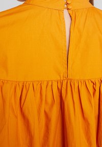 Oranssi puuvillamekko, jossa on nappisulku selässä, rypytetty helma ja sileä, teksturoitu kangas.