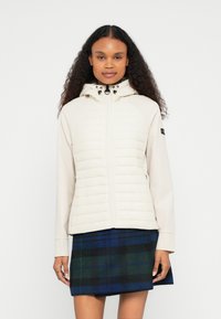 Ανοιχτό μπεζ μπουφάν με επένδυση και κουκούλα, εξοπλισμένο με φερμουάρ και ελαστικές λεπτομέρειες. Φορεμένο με σκουρόχρωμη καρό φούστα. Ομαλή υφή.