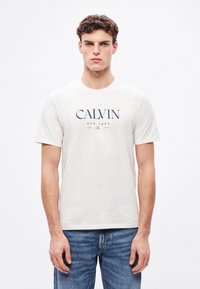 Camiseta blanca de algodón con un estampado gráfico azul que incluye el texto "CALVIN NEW YORK". El modelo la lleva con vaqueros azules de denim.