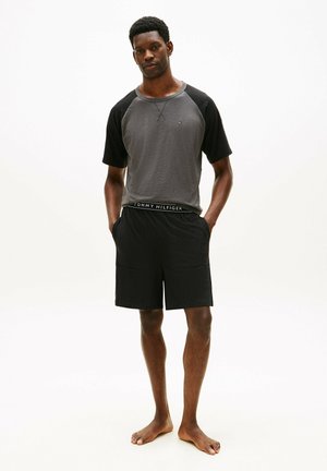 Jeune homme debout pieds nus, portant un t-shirt à manches courtes gris et noir avec un short noir, les mains dans les poches.