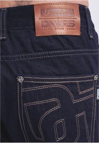 Tummansiniset farkut, joissa on nahkainen etiketti, jossa lukee "Denim by DNGRS", korostettuna kontrastivärisellä vaaleanruskealla ompeleella ja metalliosilla.