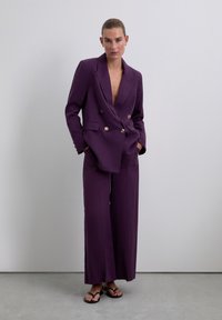 Blazer double boutonné violet avec boutons dorés, assorti à un pantalon large. Tissu lisse, coupe ajustée et design minimaliste.
