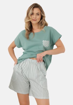 Kobieta w luźnym miętowym T-shircie z paskowaną kieszenią i dopasowanych paskowanych elastycznych szortach w talii, pozująca z jedną ręką na biodrze.