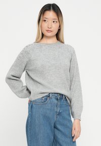 Pullover cinza feito de material suave, apresenta um decote redondo, mangas longas e detalhes decorativos de botões nos ombros. Usado com jeans azuis.