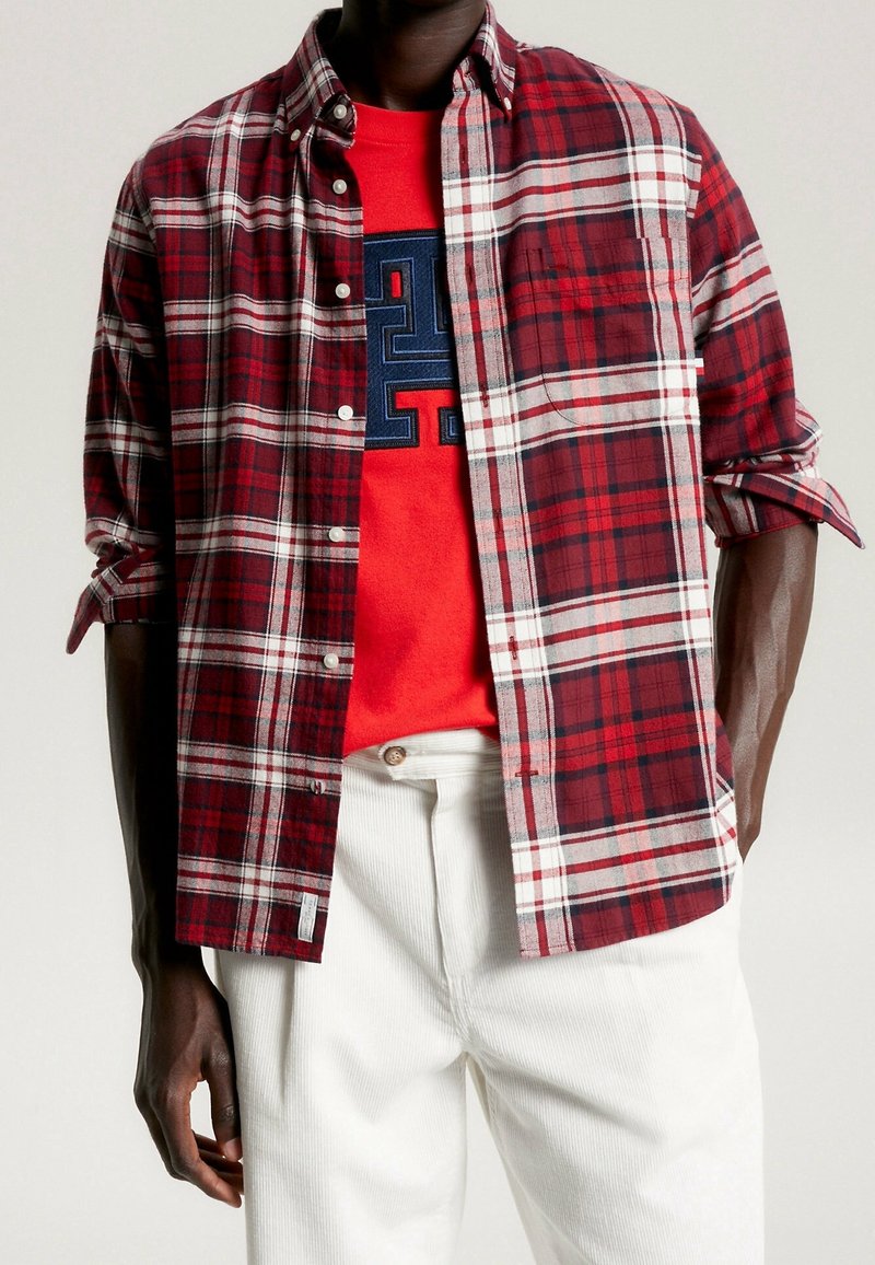 Homme portant une chemise à carreaux rouge et blanche déboutonnée sur un sweat-shirt rouge à motif graphique et un pantalon blanc sur un fond uni.