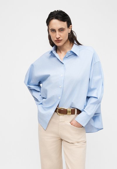 Personne aux cheveux foncés portant une chemise ample bleu clair à boutons, un pantalon beige et une ceinture marron, posant avec une main dans la poche.