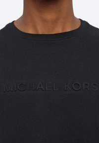 Camiseta de algodón negra con un logo en relieve y tono "MICHAEL KORS" en el pecho, con un cuello redondo clásico y una textura suave.