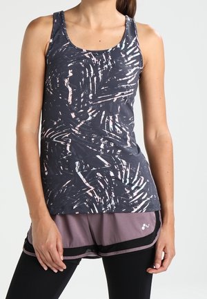 Kobieta w ciemnym, bezrękawnym topie z wzorem, lawendowych szortach z czarną lamówką i czarnych legginsach, stojąca z rozluźnionymi rękami.