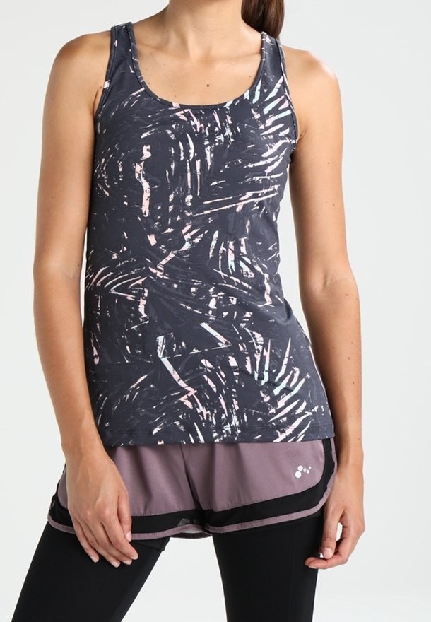 Femme portant un débardeur sombre à motifs, un short mauve avec des bordures noires et des leggings noirs, debout les mains relâchées.