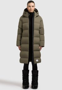 Long manteau matelassé vert olive avec une capuche, doté d'une fermeture éclair noire et d'un patch logo rectangulaire à l'ourlet ; associé à des bottes noires.