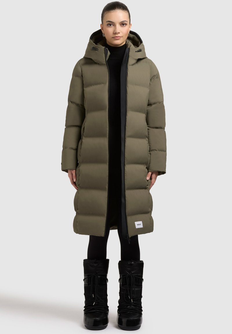 Long manteau matelassé vert olive avec une capuche, doté d'une fermeture éclair noire et d'un patch logo rectangulaire à l'ourlet ; associé à des bottes noires.