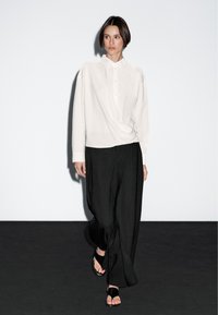 Hvid langærmet bluse med et snoet frontdesign, parret med løse sorte bukser og sorte sandaler på en mørk baggrund.