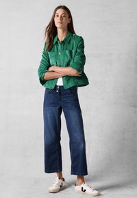 Grøn kordjakke med krave-design, parret med mørkeblå brede bukser og hvide sneakers med sorte detaljer.