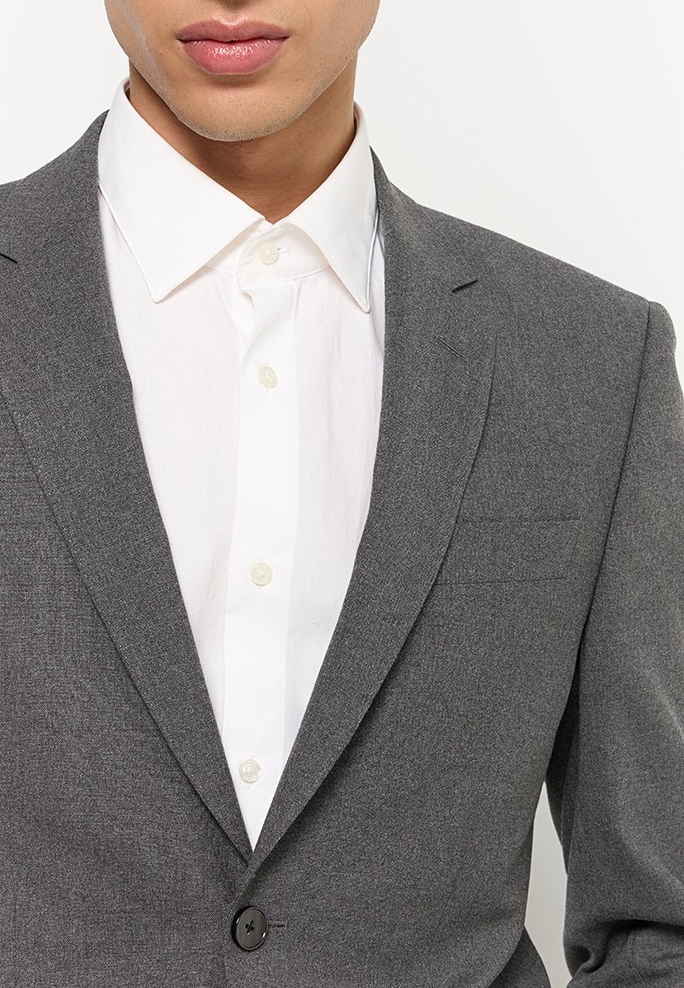Blazer gris clair avec une finition texturée, fermeture à un bouton et revers. Porté avec une chemise blanche à boutons en dessous.
