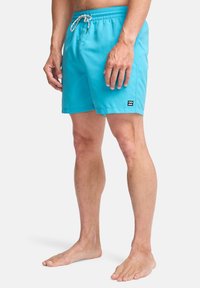 Ανοιχτό μπλε μαγιό με ελαστική μέση και κορδόνι, που διαθέτει ένα μικρό μαύρο λογότυπο στην κάτω αριστερή πλευρά.