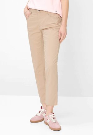 Person iført beige bukser med lige ben og sidelommer, lyserøde afslappede sneakers og en lyserød top, stående med den ene hånd i lommen.