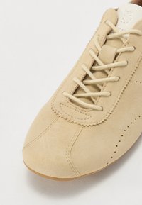 Beige semsket sko med rund tå, snøring, bølget kant og perforerte detaljer nær snøringen. Glatt lær på tungen.