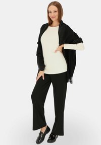 Czarny szal z frędzlami na białym swetrze. Luźne czarne spodnie i błyszczące czarne buty. Gładka dzianina i jednolite kolory.