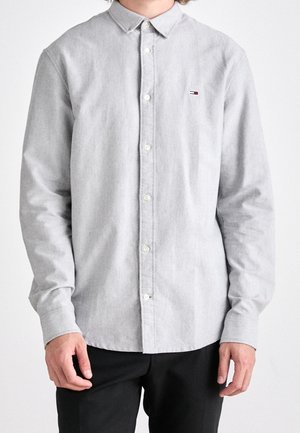 Homme portant une chemise gris clair boutonnée avec un petit logo sur la poitrine gauche et un pantalon noir sur un fond uni.