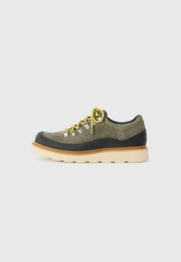 Olive green suede sneaker with black rubber trim, yellow laces, tan midsole, and off-white thick ridged outsole.