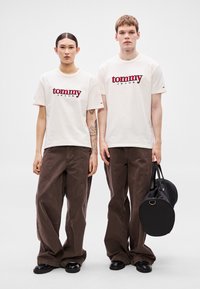 Białe koszulki z napisem "tommy JEANS" na czerwono, zestawione z szerokimi spodniami w ciemnym brązie. Jedna osoba niesie okrągłą czarną torbę z rączką.