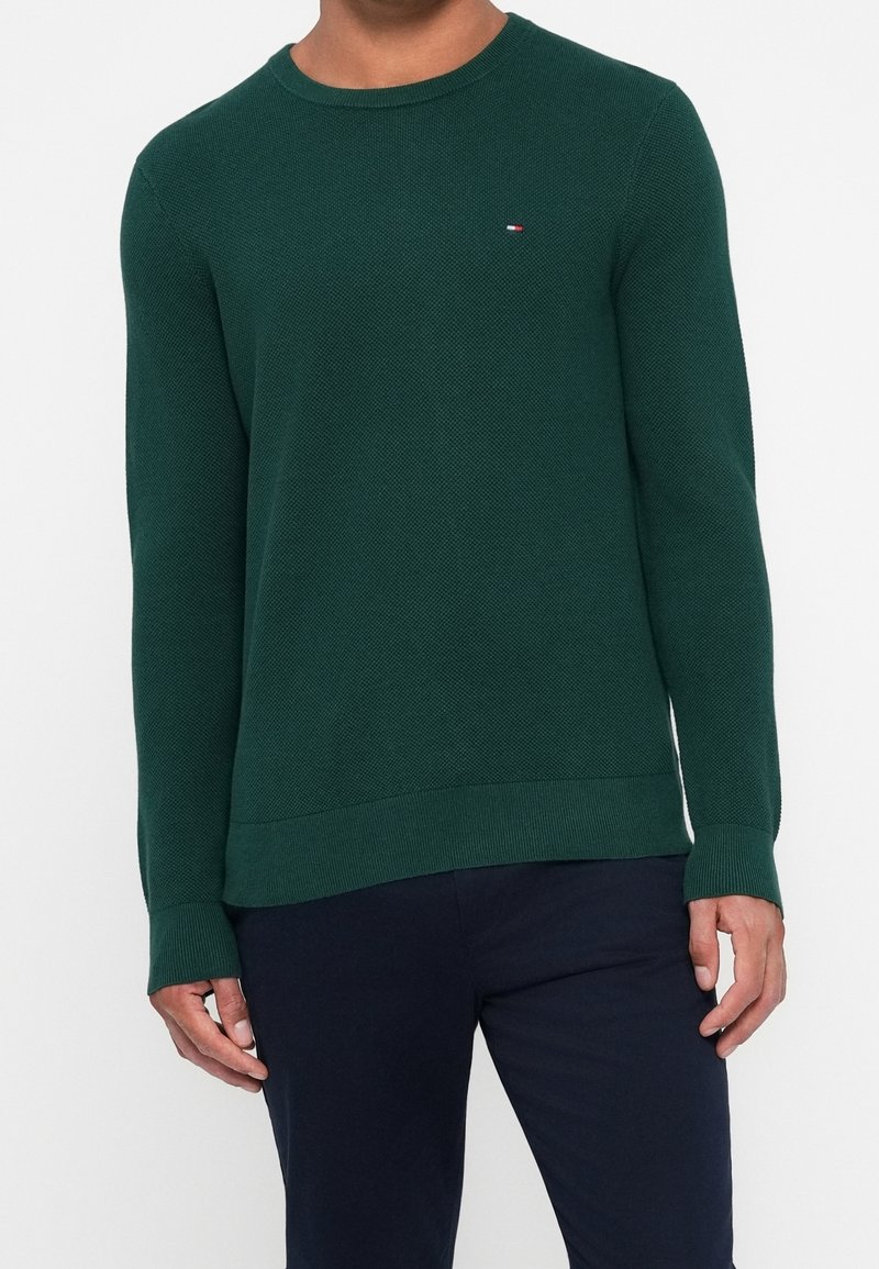 Homme portant un pull col rond texturé vert foncé avec un petit logo sur la poitrine, associé à un pantalon sombre, debout devant un fond uni.