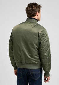 Giacca bomber verde oliva con una finitura liscia e lucida, collo e polsini a coste, e tasche laterali. Indossata sopra jeans in denim scuro.