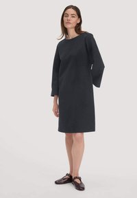 Schwarzes knielanges Kleid mit weitem U-Ausschnitt und langen, schmal zulaufenden Ärmeln. Glatter Stoff mit lockerer Passform. Getragen mit dunklen, flachen Schuhen.