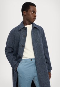 Blauwe visgraatjas met grote kraag en vijf knopen, gedragen over een crèmekleurige trui en lichtblauwe corduroybroek. Getextureerd wollen materiaal.