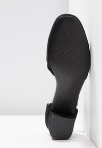 Chaussure en cuir suédé noir avec une semelle en caoutchouc texturée, dotée d'un talon en bloc bas et d'un design minimaliste, inclinée contre un fond clair.