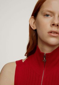Top de punto acanalado rojo con cuello alto y cierre de cremallera, que presenta cortes en los hombros. El modelo lleva un pendiente dorado y muestra un maquillaje natural.