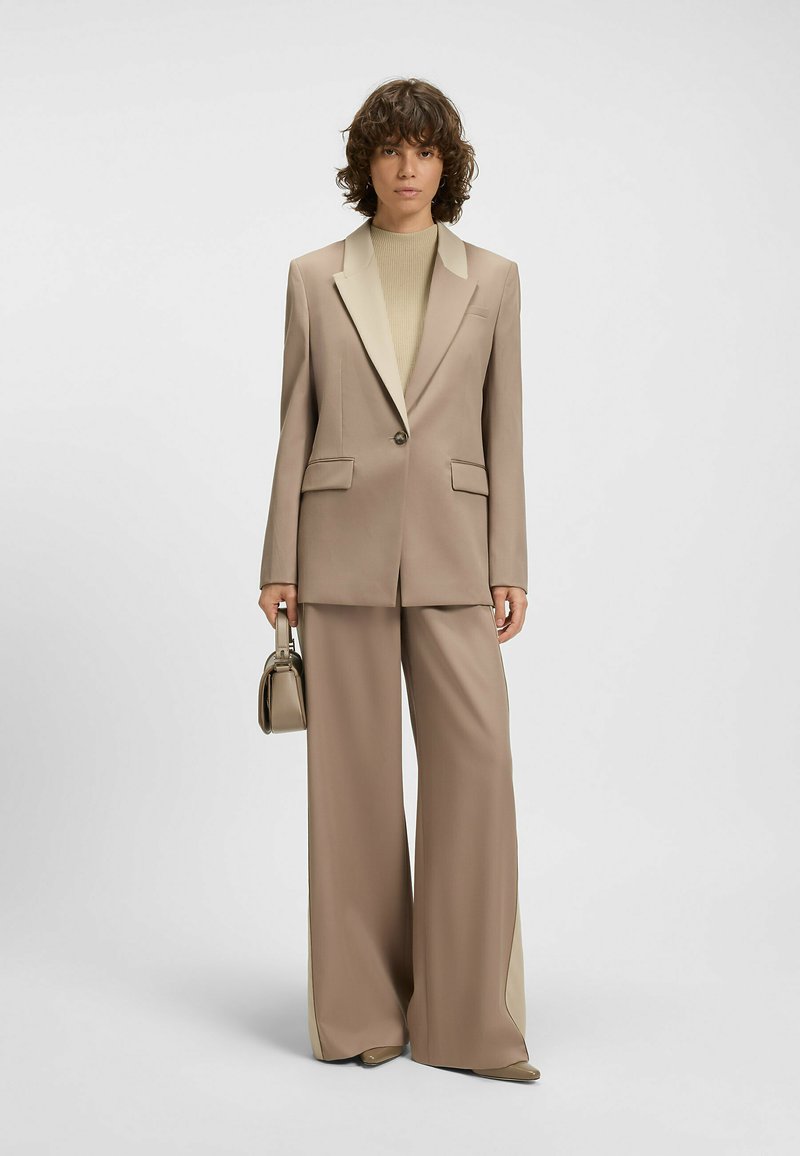 Beige pak met een op maat gemaakte blazer met een contrasterende kraag, brede broekspijpen en een gebreide top met structuur. Gecombineerd met een kleine handtas.